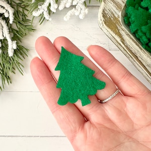 May include: A small, green felt Christmas tree held in a hand. The tree is a simple, stylized shape. Other green felt trees are in a wooden bowl. Decorative greenery with red berries and white accents are in the background.