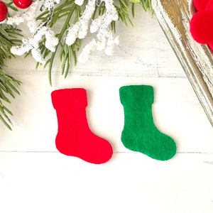 May include: Two felt Christmas stockings, one red and one green, are displayed on a white wooden surface. The stockings are small and have a classic design. Festive holiday decor.