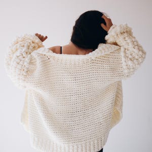 May include: A cream-coloured, oversized knitted cardigan with large, textured sleeves. The chunky knitwear has a relaxed fit and a wide, open neckline. The back view shows the garment's full length and the texture of the yarn.