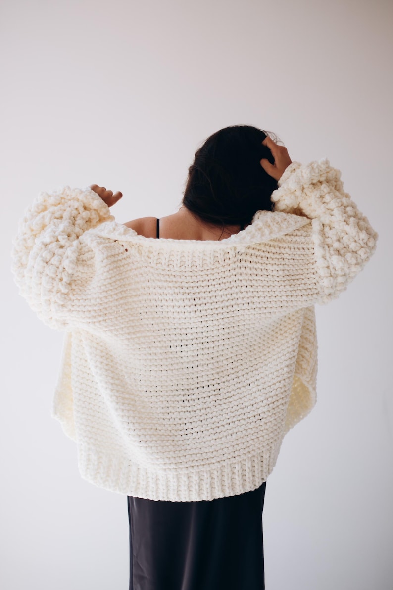 May include: A cream-coloured, oversized knitted cardigan with large, textured sleeves. The chunky knitwear has a relaxed fit and a wide, open neckline. The back view shows the garment's full length and the texture of the yarn.