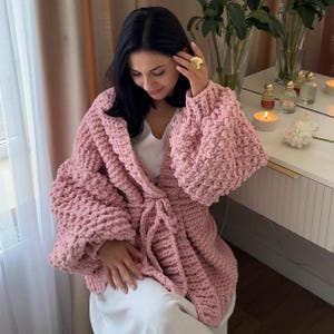 May include: A chunky knit cardigan in a soft pastel pink hue, featuring an open front design with a matching tie belt, wide sleeves, and a thick, textured yarn construction worn over a simple white garment with a gold heart-shaped ring.