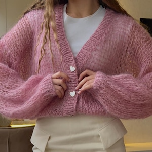 May include: A light pink, open-weave cardigan with a V-neck and long sleeves. The cardigan has two white heart-shaped buttons. The garment is made of a soft, fuzzy material.