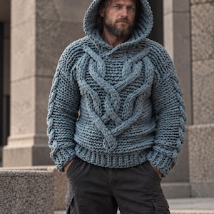 May include: A chunky knit hooded sweater in a dusty blue color featuring a prominent braided cable pattern on the front and sleeves, long sleeves with ribbed cuffs, and a thick ribbed hem, paired with dark gray cargo pants.
