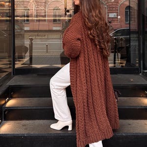 May include: A long, chunky knit cardigan in a deep brown color featuring a vertical cable knit pattern. The garment is worn over white trousers and white heeled ankle boots. The cardigan has long sleeves and reaches below the knee in length.