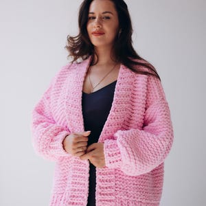 May include: A light pink, chunky knit cardigan with a shawl collar. The open-front sweater has long sleeves and a relaxed fit. The cardigan is worn over a dark blue dress. The background is a plain white.