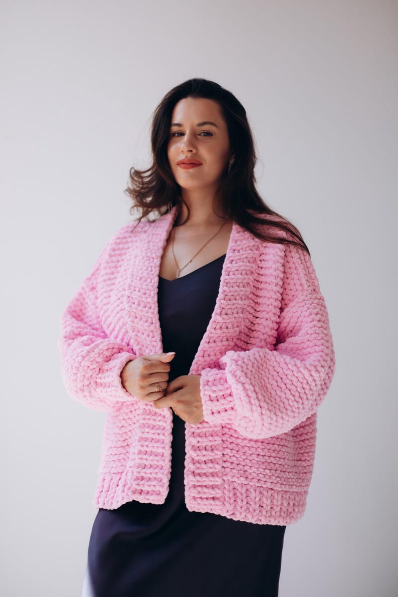 May include: A light pink, chunky knit cardigan with a shawl collar. The open-front sweater has long sleeves and a relaxed fit. The cardigan is worn over a dark blue dress. The background is a plain white.