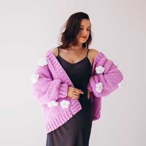 May include: A lilac chunky knit cardigan with white flower appliqu&eacute;s. The open-front sweater has long sleeves and is worn over a black slip dress. The cardigan is a statement piece, perfect for adding a touch of whimsy to any outfit.