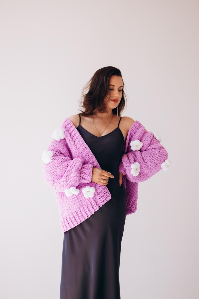 May include: A lilac chunky knit cardigan with white flower appliqu&eacute;s. The open-front sweater has long sleeves and is worn over a black slip dress. The cardigan is a statement piece, perfect for adding a touch of whimsy to any outfit.