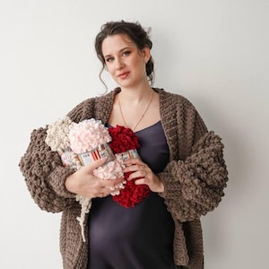 Peut inclure: Une femme porte un cardigan en tricot marron épais et tient trois pelotes de laine Alize crème, rose et rouge. La laine a un aspect texturé et bouclé, adapté aux projets de tricot ou de crochet. Le cardigan a de grandes manches bouffantes.
