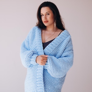 May include: A pale blue, chunky knit cardigan. The open-front sweater has long sleeves and a relaxed fit. The soft, textured knitwear is a solid colour. The cardigan is worn over a dark top and a long necklace.