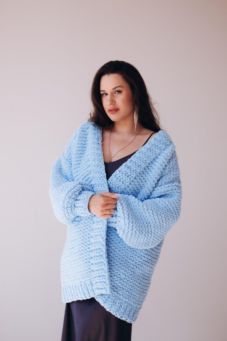 May include: A pale blue, chunky knit cardigan. The open-front sweater has long sleeves and a relaxed fit. The soft, textured knitwear is a solid colour. The cardigan is worn over a dark top and a long necklace.