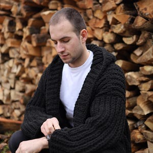 May include: A chunky black knit cardigan with a hood and long sleeves, worn over a plain white t-shirt. The cardigan features a thick, ribbed texture throughout the garment, providing a heavy and warm appearance with a wide collar and open front.