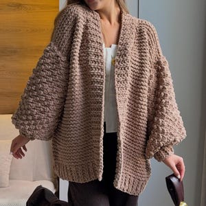 May include: A chunky, beige, open-front knit cardigan featuring a thick, textured stitch pattern, long voluminous sleeves with ribbed cuffs, and a wide ribbed hem. The garment is worn over a white top and dark brown trousers, paired with a dark brown handbag.