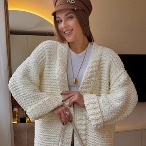 May include: Cream-colored, chunky knit cardigan with long sleeves. The open-front sweater has a textured pattern and a relaxed fit. The model is wearing a white top and a brown cap. A gold pendant necklace is visible.