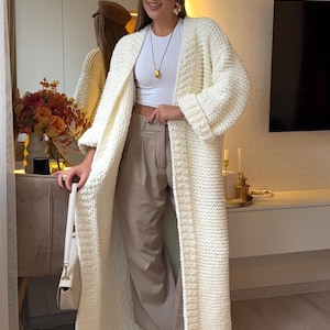 以下が含まれることがあります： クリーム色のロング丈、分厚いニットカーディガン。白いトップスとベージュのパンツの上にカーディガンを着用。白いハンドバッグと茶色のキャップも見える。カーディガンは手作りのようで、スタイリッシュなアウターです。