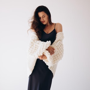 May include: A woman wearing a cream-coloured, chunky knit cardigan over a dark navy slip dress. The cardigan has a textured, oversized design. The woman has long, dark hair and is standing against a white background.