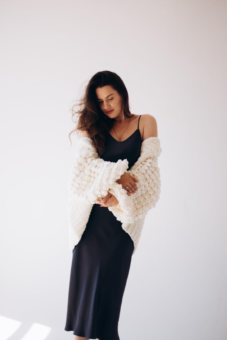 May include: A woman wearing a cream-coloured, chunky knit cardigan over a dark navy slip dress. The cardigan has a textured, oversized design. The woman has long, dark hair and is standing against a white background.