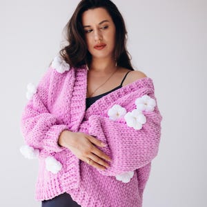 May include: A light pink, chunky knit cardigan decorated with white floral accents. The open-front sweater is made of a thick, soft yarn and has a relaxed fit. The cardigan is a cosy and stylish outerwear option.