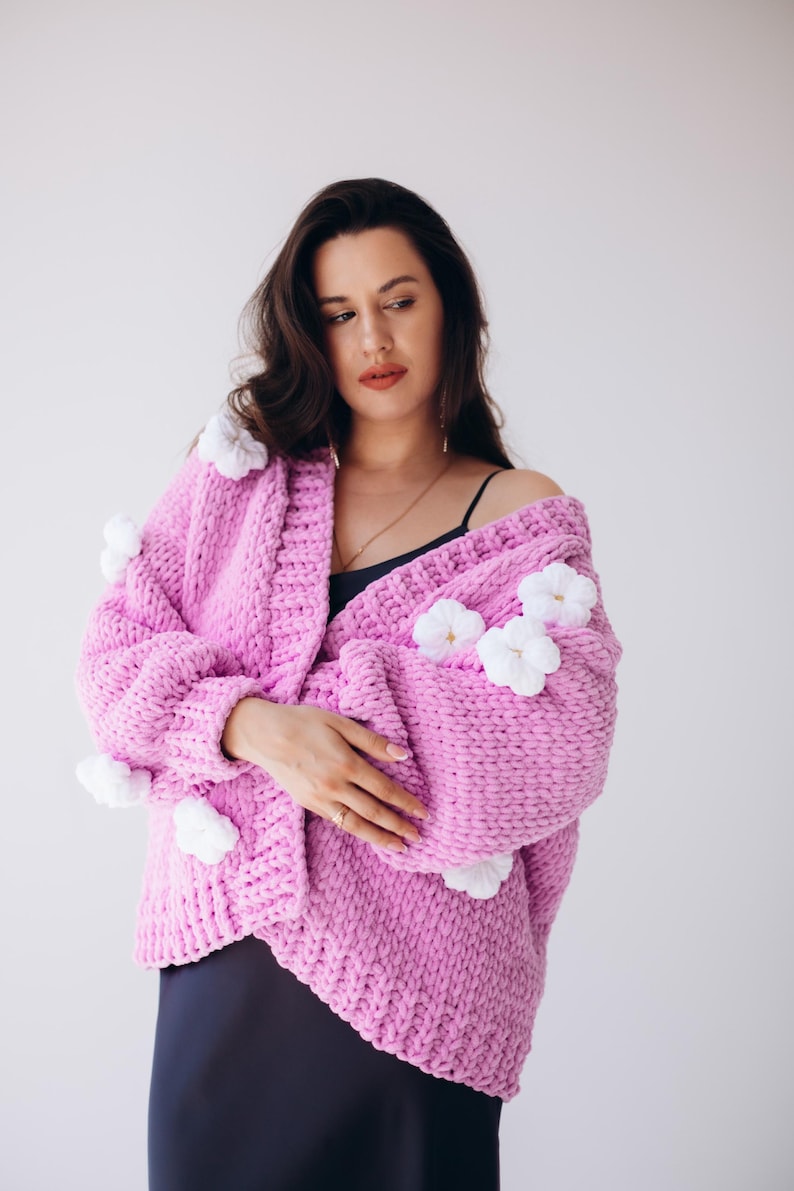 May include: A light pink, chunky knit cardigan decorated with white floral accents. The open-front sweater is made of a thick, soft yarn and has a relaxed fit. The cardigan is a cosy and stylish outerwear option.