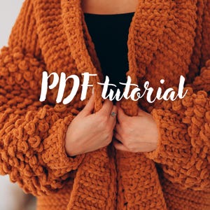 May include: Close-up of a chunky knit orange cardigan. The cardigan has a thick, textured appearance with large, looped stitches. The words "PDF tutorial" are written across the front in a cursive font.
