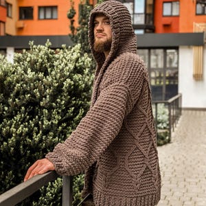 May include: A brown hooded cardigan with a chunky knit design. The sweater features a diamond pattern on the front and a ribbed hem and cuffs. The model is wearing the cardigan outdoors.