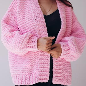 May include: A pale pink, chunky knit cardigan with a shawl collar. The open-front sweater has long sleeves and a textured, ribbed design. The cardigan is made of a thick, plush material, ideal for layering.