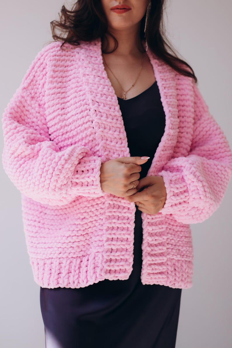 May include: A pale pink, chunky knit cardigan with a shawl collar. The open-front sweater has long sleeves and a textured, ribbed design. The cardigan is made of a thick, plush material, ideal for layering.