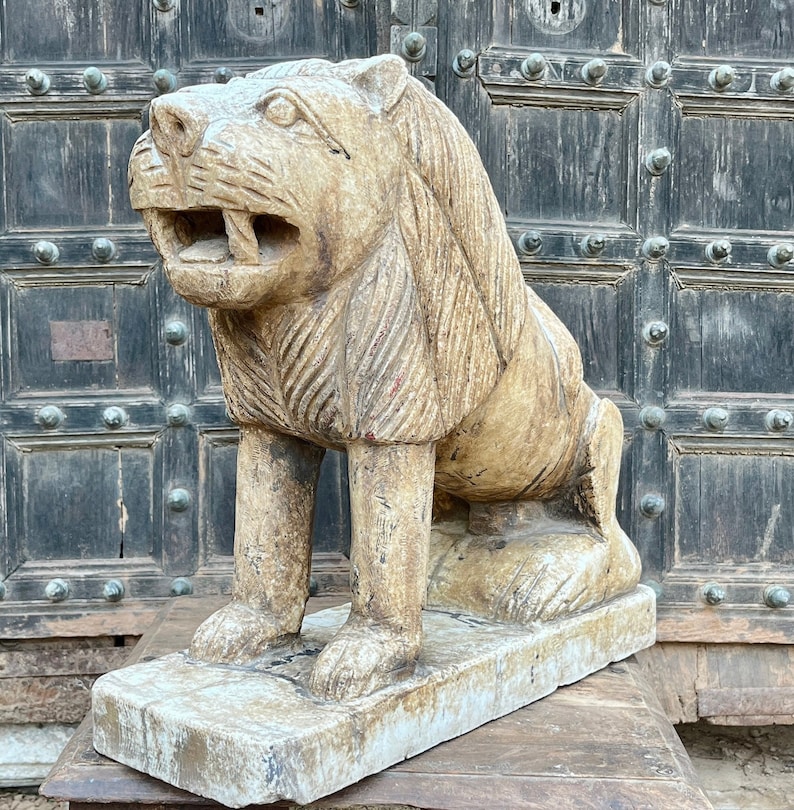 Puede incluir: Una estatua de le&oacute;n de piedra tallada con una melena detallada y una boca abierta de par en par. La estatua est&aacute; pintada de un color beige claro y se encuentra sobre una base de piedra blanca.