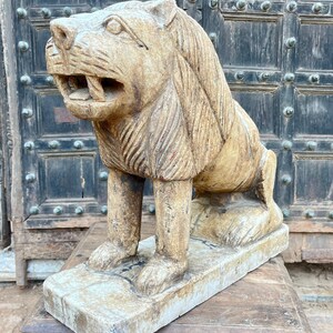 Puede incluir: Una estatua de le&oacute;n de piedra tallada con una melena detallada y una boca ligeramente abierta. La estatua est&aacute; sentada sobre una base de piedra blanca y est&aacute; en una posici&oacute;n ligeramente agachada.