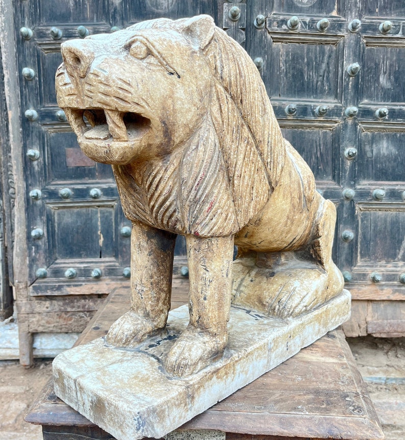 Puede incluir: Una estatua de le&oacute;n de piedra tallada con una melena detallada y una boca ligeramente abierta. La estatua est&aacute; sentada sobre una base de piedra blanca y est&aacute; en una posici&oacute;n ligeramente agachada.