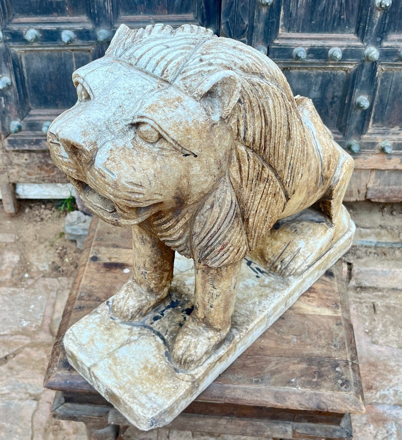 Puede incluir: Una estatua de le&oacute;n de piedra tallada con un acabado blanco desgastado. El le&oacute;n est&aacute; de pie sobre una base rectangular y tiene una melena y una cara detalladas.