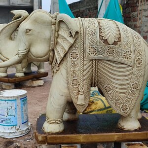 Puede incluir: Una estatua de elefante de m&aacute;rmol blanco con tallas intrincadas y un pa&ntilde;o decorativo drapeado sobre su espalda. El elefante est&aacute; de pie sobre una base de madera.