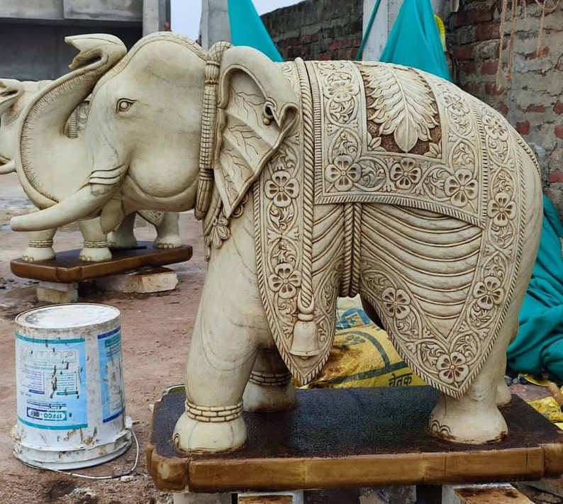 Puede incluir: Una estatua de elefante de m&aacute;rmol blanco con tallas intrincadas y un pa&ntilde;o decorativo drapeado sobre su espalda. El elefante est&aacute; de pie sobre una base de madera.