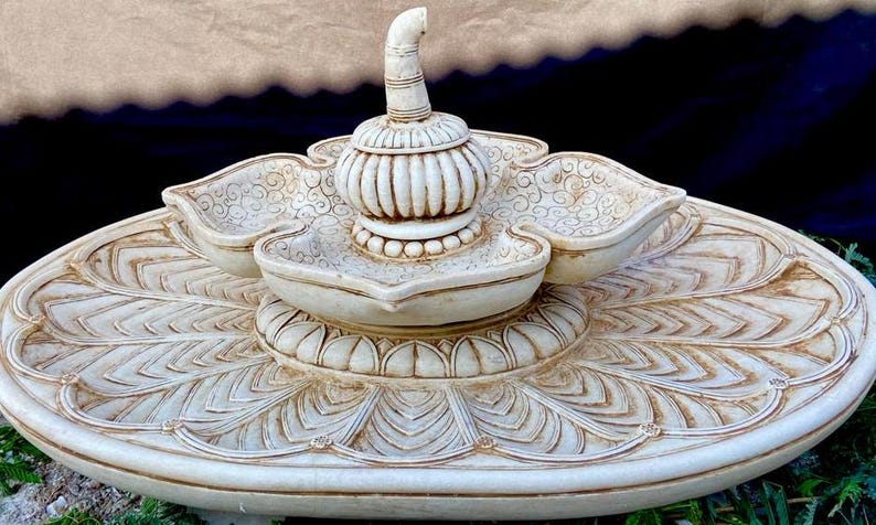 May include: A white ceramic fountain with a tiered design featuring intricate carvings. The fountain has a central spout and three smaller bowls, all decorated with floral patterns.