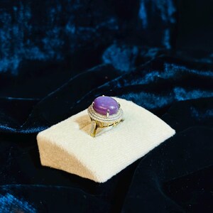 Puede incluir: Un anillo con una gran piedra preciosa ovalada de color púrpura rodeada de piedras preciosas más pequeñas y transparentes. El anillo está en un expositor blanco.