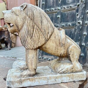 Puede incluir: Una estatua de le&oacute;n de madera tallada con un acabado blanco y marr&oacute;n claro. El le&oacute;n est&aacute; de pie sobre una base rectangular y tiene una melena y una cara detalladas.
