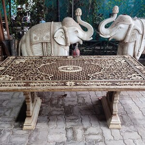 Puede incluir: Una mesa de madera grande, tallada de forma intrincada con un dise&ntilde;o marr&oacute;n y blanco. La mesa tiene una tapa rectangular y dos patas a cada lado. La mesa est&aacute; delante de dos estatuas de elefantes blancos.