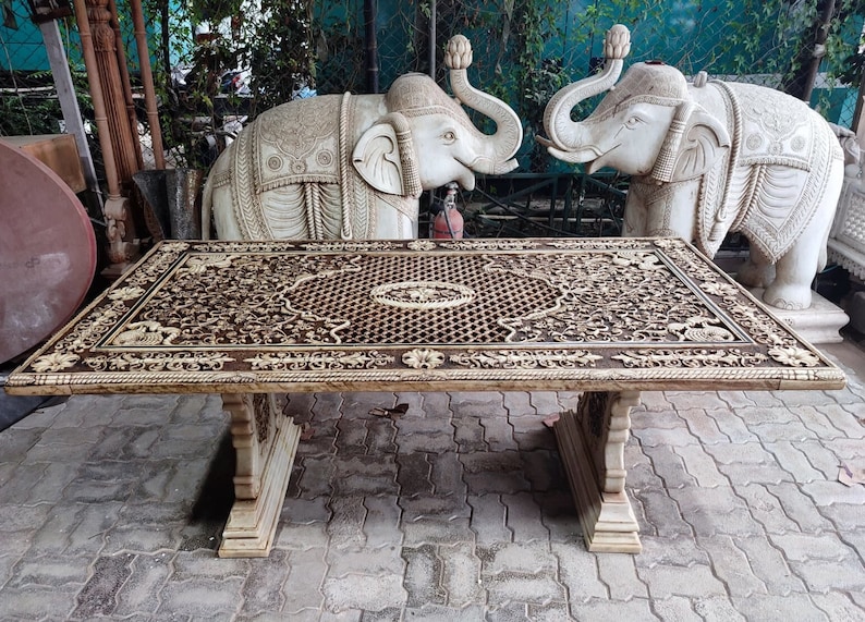 Puede incluir: Una mesa de madera grande, tallada de forma intrincada con un dise&ntilde;o marr&oacute;n y blanco. La mesa tiene una tapa rectangular y dos patas a cada lado. La mesa est&aacute; delante de dos estatuas de elefantes blancos.