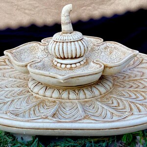 May include: A white ceramic fountain with a lotus flower design. The fountain has three tiers and a central spout. The fountain is decorated with intricate carvings.
