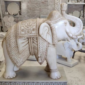 Puede incluir: Una estatua de elefante de m&aacute;rmol blanco con tallas intrincadas y una silla de montar decorativa. El elefante est&aacute; de pie sobre una base con un borde decorativo.