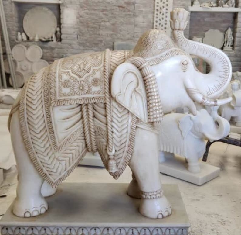 Puede incluir: Una estatua de elefante de m&aacute;rmol blanco con tallas intrincadas y una silla de montar decorativa. El elefante est&aacute; de pie sobre una base con un borde decorativo.