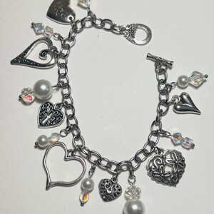 May include: A silver-toned charm bracelet featuring various heart-shaped charms, including one engraved with the text I love you, a cross charm, and a floral heart charm. The bracelet is accented with white faux pearl beads and clear faceted crystals.