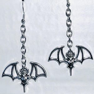 May include: A pair of silver dangle earrings featuring a bat design. Each earring has a silver chain and a bat charm with spread wings.