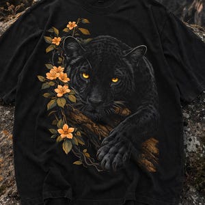 Puede incluir: Camiseta negra con una ilustración detallada de una pantera negra con ojos amarillos, descansando sobre una rama. Flores naranjas y hojas verdes en el lado izquierdo. La camiseta tiene un aspecto vintage y desgastado.