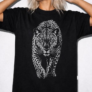 May include: A black short-sleeved t-shirt featuring a large, detailed white graphic print of a leopard walking forward. The leopard is depicted with realistic spots and intense eyes, centered on the chest of the garment. The shirt has a crew neckline.
