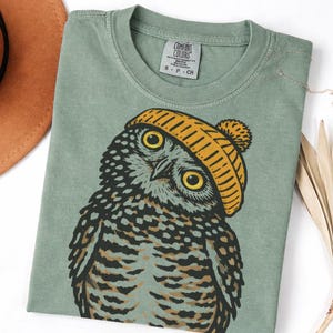 May include: Sage green Comfort Colors t-shirt featuring a detailed illustration of an owl wearing a yellow beanie. The owl has large yellow eyes and is set against a neutral background. The shirt has a classic crew neck and short sleeves.
