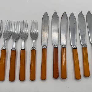 Pode incluir: Um conjunto de talheres vintage, incluindo garfos e facas. Os garfos têm dentes prateados e cabos laranja-castanhos. As facas têm lâminas prateadas e cabos laranja-castanhos a condizer. Os talheres estão dispostos numa superfície branca.