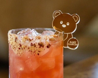 May include: A pink cocktail with ice cubes in a clear glass with a sugared rim. The glass has a clear plastic bear topper with the text "BABY GARCIA" on it.