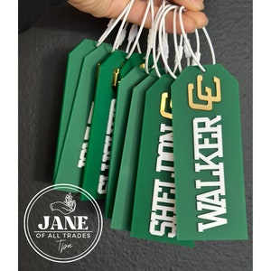 May include: A stack of green gift tags with white and gold lettering. The tags are tied together with white string. The text on the tags reads "SHELTON" and "WALKER".