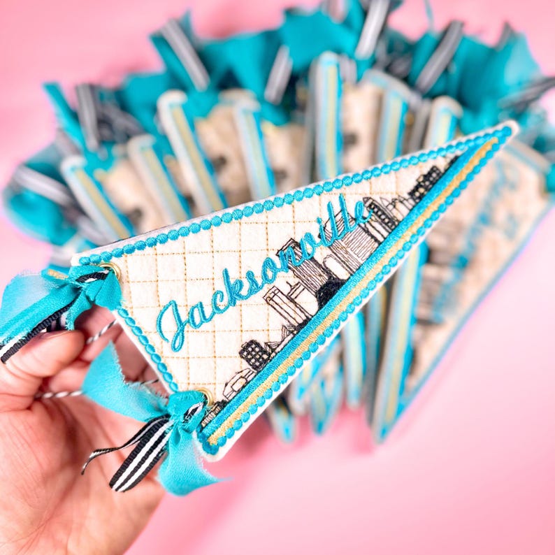 May include: A cream-colored pennant with the word "Jacksonville" embroidered in teal script, along with a skyline illustration. The pennant is trimmed with teal and gold stitching and has a teal bow with black and white striped ribbon.